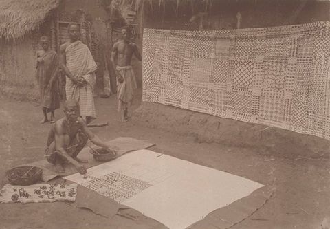 Sepia photograph. A group of four men are documented in an outdoor setting. Behind them is a partial view of a simple, single storey building with a straw roof. To their left, a section of wall is covered by a textile featuring an intricate geometric pattern. In the foreground, a crouching man is seen painting a pattern onto another sheet of fabric laid out flat on the ground.