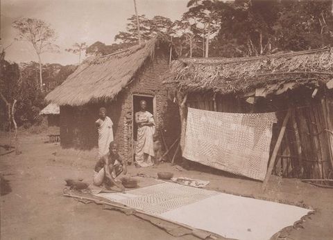 Sepia photograph. Two people stand in front of a small, rustic building with a straw roof, while a third crouches in the foreground before a partially painted textile laid out flat on the floor. To their left, another textile featuring a geometric pattern is slung over a washing line. 