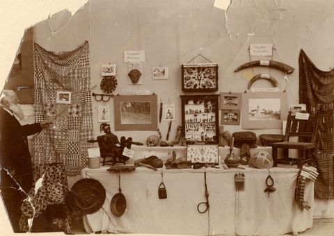 Sepia photograph. In this document of an old photograph, a group of African objects and artefacts are arranged into a display across a wall and onto a table covered by a white tablecloth. To the left, a white man seen side on points towards one of the objects with a long stick. The original, sepia photograph shows signs of damage, with ripped edges and creases.