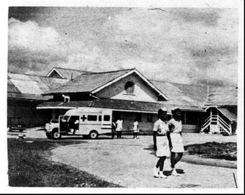 Black and white photograph. A wide, curved road leads towards a building in the distance, which has a slanted, slated roof and a round window in the centre. In front of the building a white van with a cross on it has doors swung open, and two people dressed in white seem to load something out of the back. In the foreground, two black women wearing white, medical uniforms walk side-by-side around the curved bend of the road.