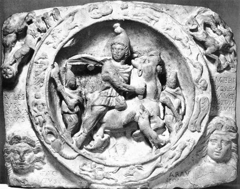 Black and white photograph. A low-relief stone sculpture depicting a young person riding on an animal’s back, contained within a circular shape. Around the circle, excerpts of Latin text are just visible, along with further carvings depicting faces and mythological animals.