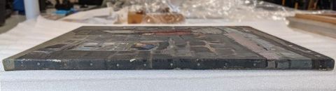 Colour photograph. The view of a painting depicting grey brickwork, laid out onto a table, seen from a side-on view. A row of nails holding the canvas to the frame can be seen. In the distance, art packing materials are just visible. On the right, part of another artwork or frame can be seen.