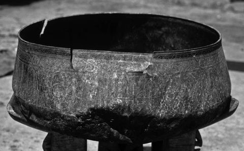 Black and white photograph. Documentation of a wide, circular vessel seen close up, with its inner chamber just visible, resting on a stone or wood base. Its outer surface is encircled with a series of intricate, geometric and circular patterns. The vessel has an aged appearance, with visible cracks.