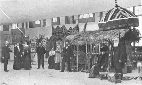 Black and white photograph. In this slightly out of focus photograph we see a row of smartly dressed white people standing or sitting within a large interior space. Behind them are a series of objects on display. On the left, a white arrow on the surface of the photograph points towards an object in the far distance.