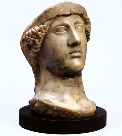 Colour photograph. A document of a carved stone head and neck resting on a black circular base. Curly hair partially covers the ears, while the rim of a cap can be seen on top. The top part of the head appears to be missing, while at the base of the neck, the rough texture of the stone is just visible.