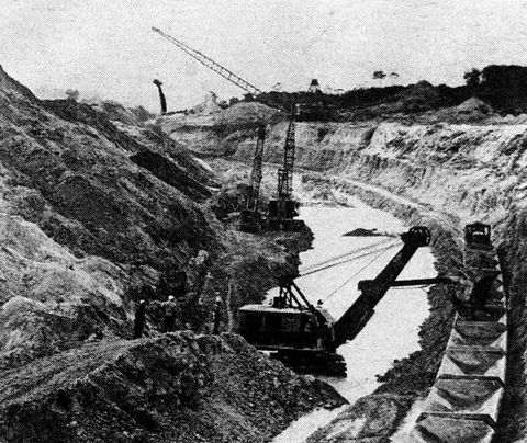 Black and white photograph. This grainy, halftone photograph depicts craggy rocks leading into a quarry, where construction tools carry out their work. On the left, a small gathering of people can be seen from behind, looking into the construction site.