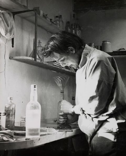 Black and white photograph. A document of a person leaning over a worktop with an implement clasped by two hands, one over the other. They are surrounded by a variety of bottles, jars, and containers on the worktop and shelves around them.