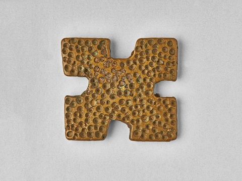 Colour photograph. A gold-toned metal object resembling a square, with 4 cut-out u shapes between the lines where two corners meet. The surface of the object has been lightly punctured with a series of circular indents to give it a mottled and uneven texture. 