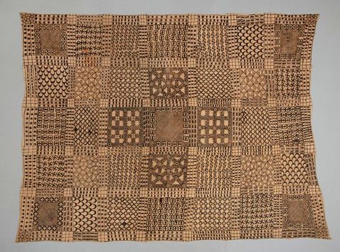 Photograph. A pale brown textile features an intricate, detailed design. The pattern of the textile is arranged into a grid formation, with squares resembling tiles, and each square ‘tile’ features its own unique geometric pattern. Rows of three intersecting lines run between the tiles, and in these are more elements of decorative pattern.