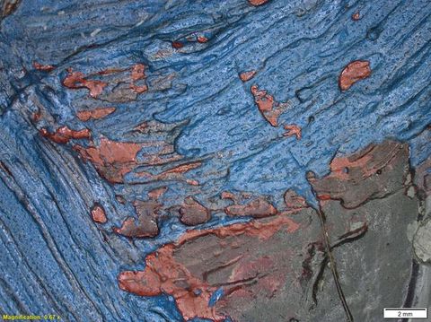 Colour photograph. A close-up, detailed study of a textural area of paint, featuring shades of blue, red, and brown, with a glossy appearance. On the lower left yellow text reads, ‘Magnification: 0.67 x’. On the right, a small white rectangle bears the text ‘2 mm’.