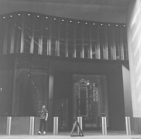 Black and white photograph. The façade of a modern building is lined by a series of silver bollards. A sign before the building reads, ‘London Mithraeum’. A casually dressed man in a patterned t-shirt and white trainers can be seen walking out of the building. Lights within the building make fragments of the building interior just visible.