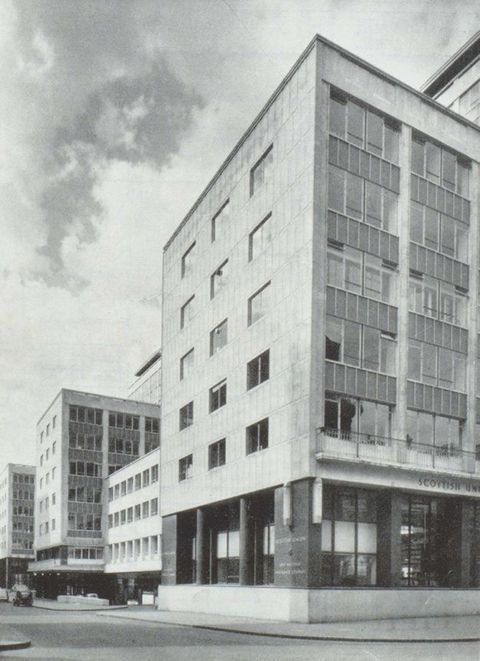 Black and white photograph. Documentation of a view down a street with a row of modern multi-storey buildings within an urban setting. On the façade of the nearest building, the word ‘Scottish’ can just be read. One black car can be seen further down the street.
