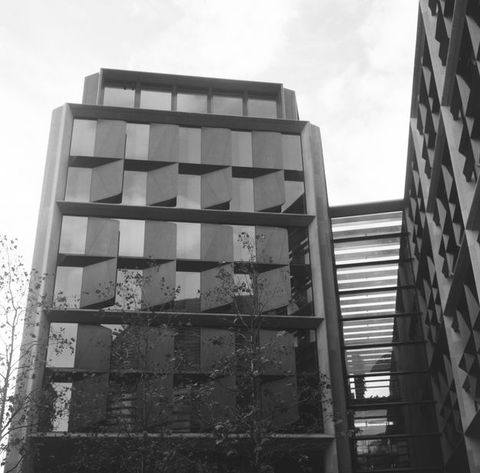 Black and white photograph. A view of modern buildings seen from below to form a series of sharp angles against the sky. A row of three almost bare trees reaches up in front of the building facing us.
