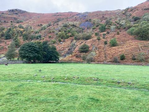 Colour photograph. Document of a rural scene showing an open, flat area of green grass divided horizontally by a slim stream. In the distance is the face of steep mountainside that leans up towards a small band of pale blue sky.