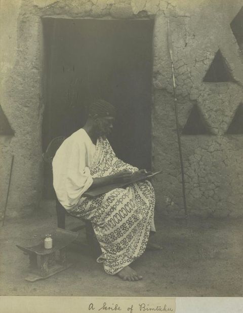 Black and white photograph. A man sits on a chair in front of an open doorway. He is dressed in pale fabric with an ornate pattern. The man holds out a writing surface and appears to be writing onto it. Next to him is a small stool containing a pot of ink. Below him, a passage of handwritten text reads, ‘A scribe of Bimtuku.’