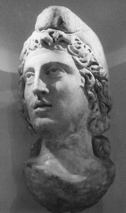 Black and white photograph. A document of a stone head statue, hung onto a white wall. The carved figure has curly hair covering their ears and wears a cap partially covering the top of their head. Their eyes look upwards and lips are slightly parted.