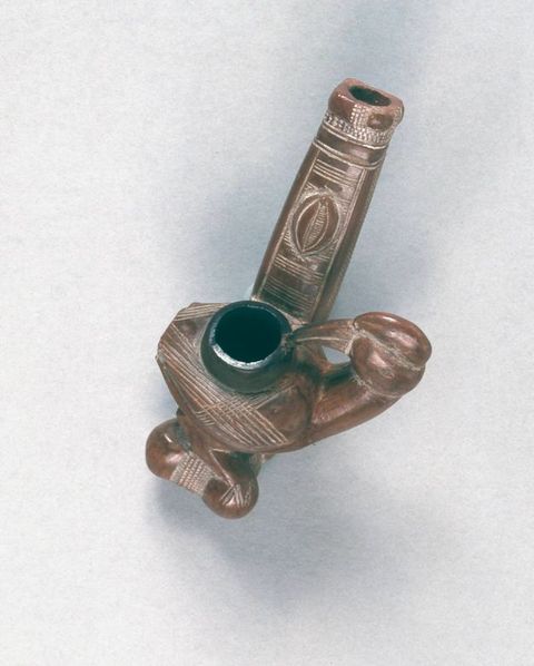 Colour photograph. A carved wooden utensil or pipe in the shape of a bird. In the centre of its back is an open hole that runs upwards and forms a pipe out through the long, straight tail. Its head turns inwards as if looking towards the hole. A series of decorative patterns have been scored into the surface of the bird.