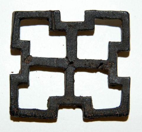 Colour photograph. An angular metal object resembling an open 2 x 2 grid is documented against a plain white background. The object shows signs of wear and tear, with elements of brown rust and cracks deeply embedded into its surface. 